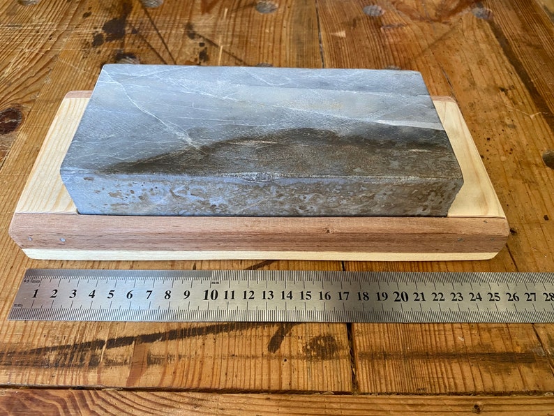 Puede incluir: Una piedra de afilar gris con una base de madera. La piedra mide aproximadamente 33 cm de largo.