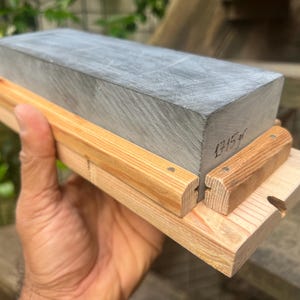 May include: A rectangular gray sharpening stone mounted on a light-colored wooden base with two side rails. The stone features the handwritten text 1715gr on its side, indicating its weight in grams. The base is constructed from light wood.