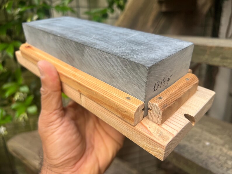 May include: A rectangular gray sharpening stone mounted on a light-colored wooden base with two side rails. The stone features the handwritten text 1715gr on its side, indicating its weight in grams. The base is constructed from light wood.