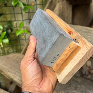 May include: A rectangular sharpening stone with a grey, textured surface mounted on a light-colored wooden base. The side of the stone features the handwritten text 1315 gr, indicating its weight in grams, with visible wood grain on the base.