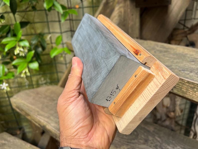 May include: A rectangular sharpening stone with a grey, textured surface mounted on a light-colored wooden base. The side of the stone features the handwritten text 1315 gr, indicating its weight in grams, with visible wood grain on the base.