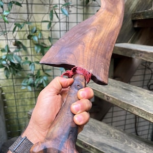 Puede incluir: Una espátula de madera hecha a mano con una hoja ancha y curvada y un acabado marrón oscuro. El mango está intrincadamente tallado y conectado a la hoja con un detalle rojo tejido. La espátula se sostiene en una mano, mostrando la veta natural de la madera.