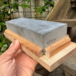 May include: A rectangular gray sharpening stone set in a light-colored wooden base. The stone features visible surface scratches and the text 1715 gr written in black ink on its side. The wooden base consists of a flat bottom piece with two side rails.