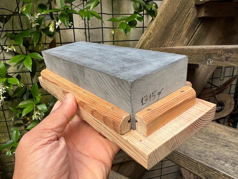 May include: A rectangular gray sharpening stone set in a light-colored wooden base. The stone features visible surface scratches and the text 1715 gr written in black ink on its side. The wooden base consists of a flat bottom piece with two side rails.