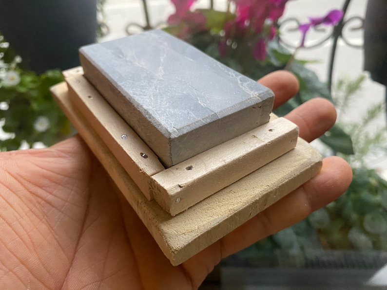 Peut inclure: Une pierre &agrave; aiguiser grise avec une base en bois. La pierre est rectangulaire et a une surface rugueuse. La base est faite de deux morceaux de bois, l'un sur l'autre. La base est tenue dans une main.