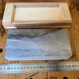 Puede incluir: Una piedra de afilar rectangular gris con una caja de madera. La piedra mide aproximadamente 25 cm de largo y 5 cm de ancho. La caja es de madera clara y tiene un borde de madera oscura.