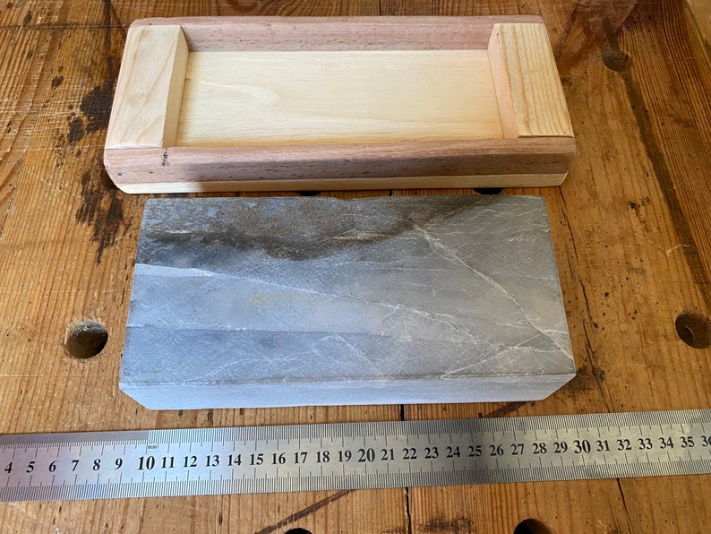 Puede incluir: Una piedra de afilar rectangular gris con una caja de madera. La piedra mide aproximadamente 25 cm de largo y 5 cm de ancho. La caja es de madera clara y tiene un borde de madera oscura.