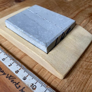 May include: A gray sharpening stone with a wooden base. The stone is rectangular and has a rough surface. The base is made of light wood and has a slightly curved shape.