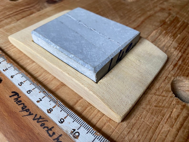May include: A gray sharpening stone with a wooden base. The stone is rectangular and has a rough surface. The base is made of light wood and has a slightly curved shape.