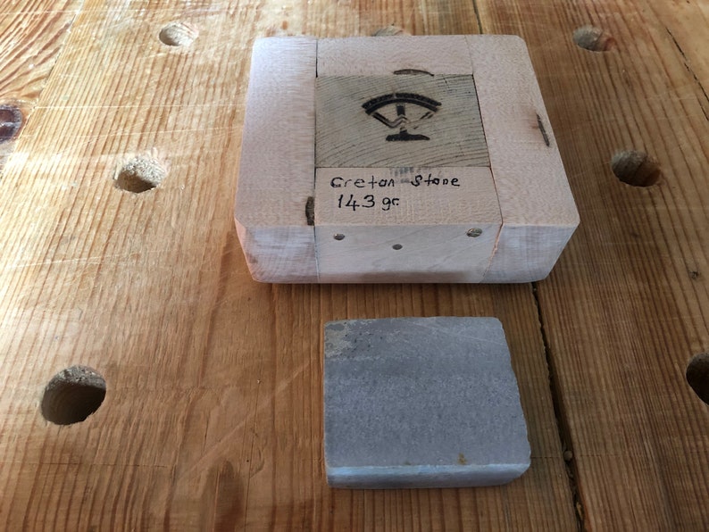 Pu&ograve; includere: Una pietra per affilare grigia su un banco da lavoro in legno. La pietra per affilare &egrave; etichettata "Gretton Stone 14.3 gr".