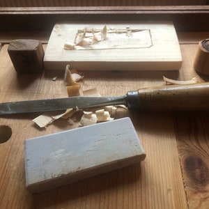 May include: A woodworking workbench with a variety of tools, including a chisel, a block of wood, a sharpening stone, and wood shavings.