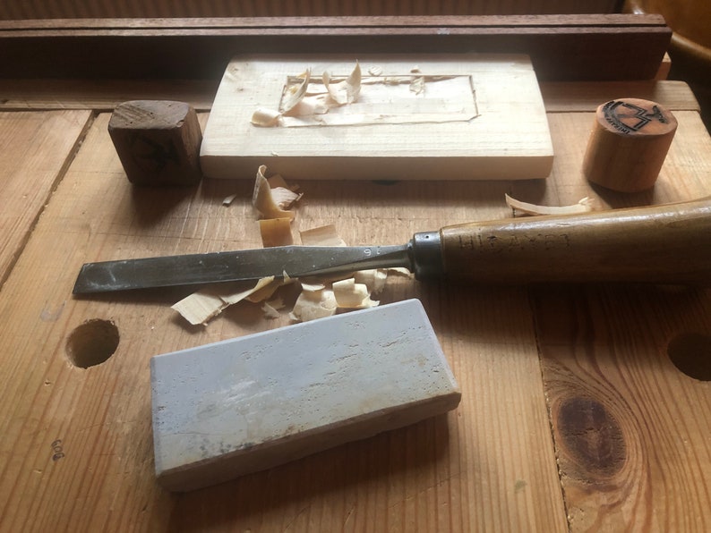 May include: A woodworking workbench with a variety of tools, including a chisel, a block of wood, a sharpening stone, and wood shavings.