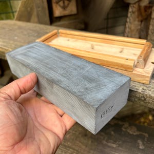 May include: A rectangular gray sharpening stone with visible surface scratches and the handwritten text 1715gr on the side, positioned next to a light-colored wooden base with raised edges designed to hold the stone securely in place.