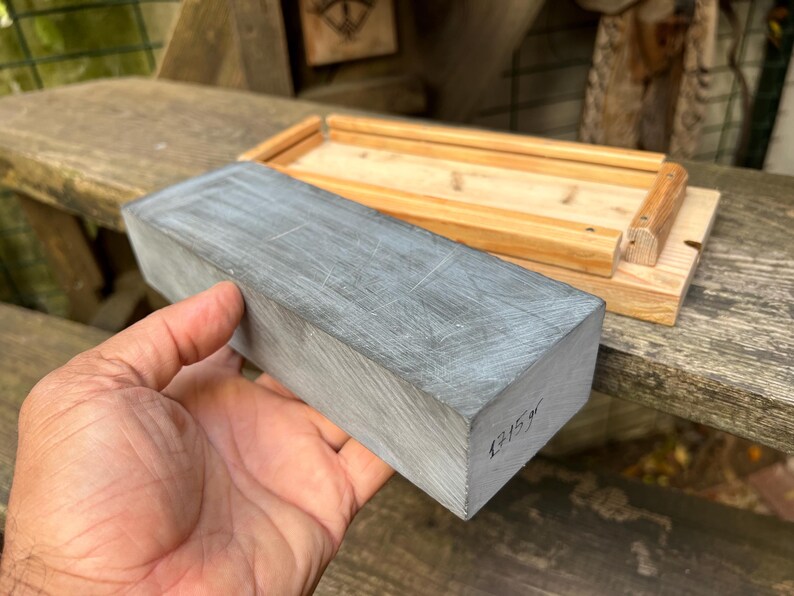 May include: A rectangular gray sharpening stone with visible surface scratches and the handwritten text 1715gr on the side, positioned next to a light-colored wooden base with raised edges designed to hold the stone securely in place.