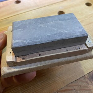 Peut inclure: Une pierre &agrave; aiguiser grise est mont&eacute;e dans un cadre en bois. Le cadre est en bois clair et a une forme rectangulaire. La pierre est maintenue en place par des vis.