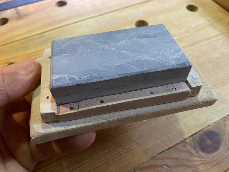 Peut inclure: Une pierre &agrave; aiguiser grise est mont&eacute;e dans un cadre en bois. Le cadre est en bois clair et a une forme rectangulaire. La pierre est maintenue en place par des vis.
