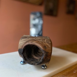May include: A wooden bowl with a natural, rustic finish. The bowl has a unique, irregular shape and a hollow center. It is mounted on three metal legs.