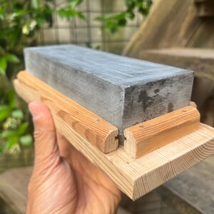 May include: A rectangular gray sharpening stone set within a light-colored wooden base. The stone features a smooth, flat top surface and is secured by two parallel wooden rails attached to the base, which has a visible wood grain texture.