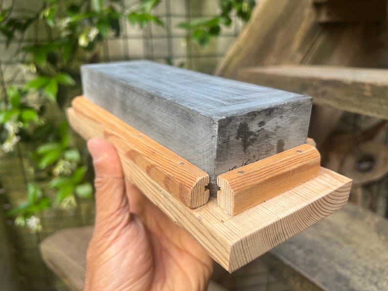 May include: A rectangular gray sharpening stone set within a light-colored wooden base. The stone features a smooth, flat top surface and is secured by two parallel wooden rails attached to the base, which has a visible wood grain texture.