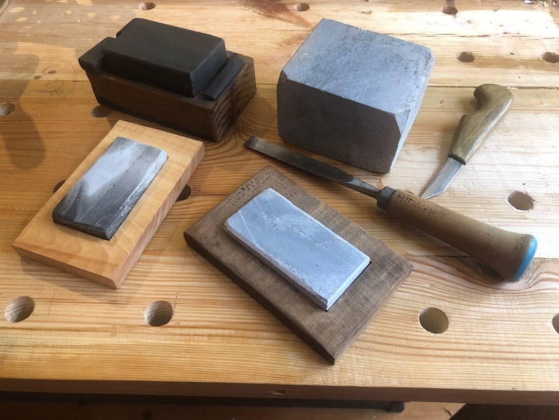 Puede incluir: Cuatro piedras de afilar, dos en marcos de madera, una en una caja de madera y una suelta. Un cincel con mango de madera con una punta azul y el texto "HITACHI" en el mango. Un cuchillo con mango de madera con un mango marr&oacute;n.