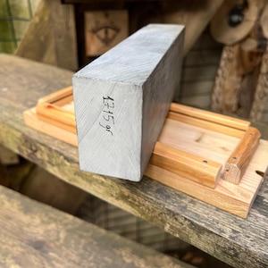 May include: A rectangular block of grey wood with a visible grain pattern, featuring the handwritten text 1715 gr on its side. The block is positioned on a light-colored wooden base with raised edges and small metal fasteners at the corners.