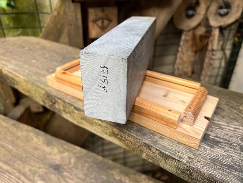 May include: A rectangular block of grey wood with a visible grain pattern, featuring the handwritten text 1715 gr on its side. The block is positioned on a light-colored wooden base with raised edges and small metal fasteners at the corners.