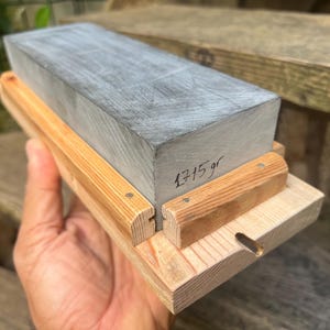 May include: A rectangular gray sharpening stone set in a light-colored wooden base with two side rails. The stone features the handwritten text 1715 gr on its side, indicating its weight in grams. The wooden base has a small circular cutout.