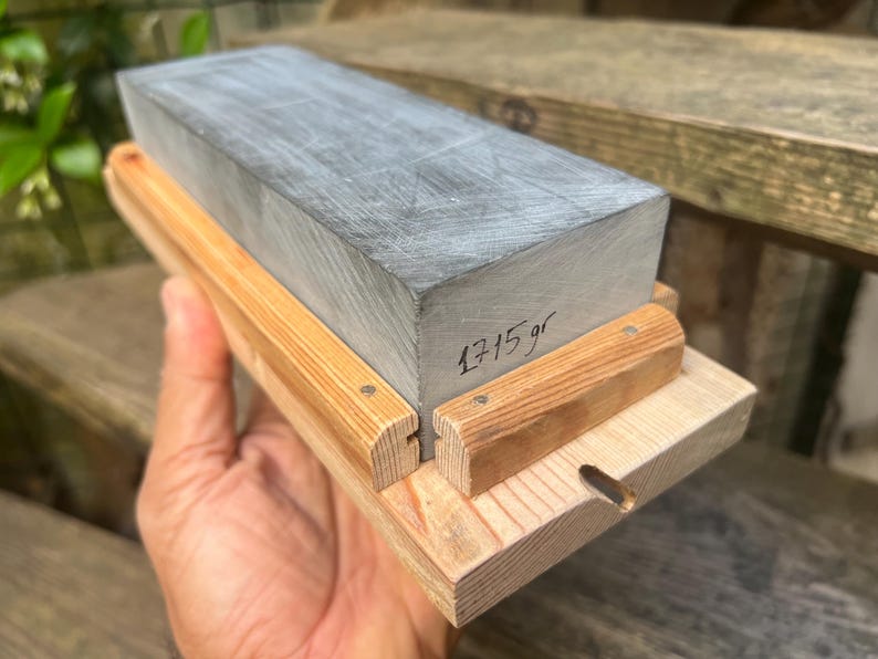 May include: A rectangular gray sharpening stone set in a light-colored wooden base with two side rails. The stone features the handwritten text 1715 gr on its side, indicating its weight in grams. The wooden base has a small circular cutout.