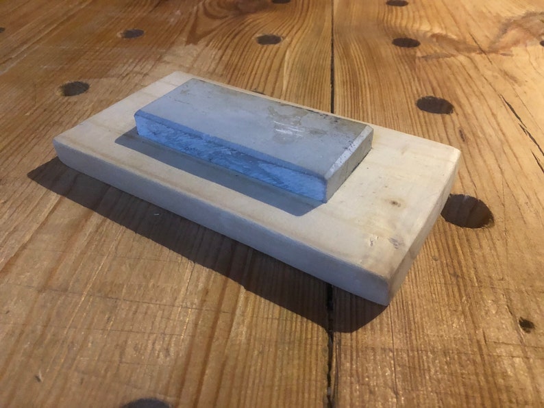 May include: A grey rectangular sharpening stone resting on a light wood base. The stone is slightly textured and has a light grey colour.