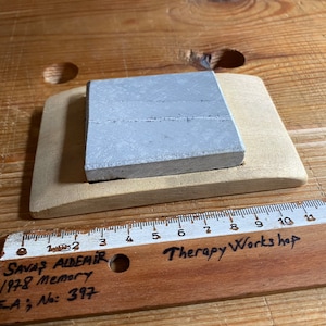May include: A gray square stone resting on a wooden base. The base is curved and has a cutout for the stone. A wooden ruler with the text "Therapy Workshop" is visible in the foreground.