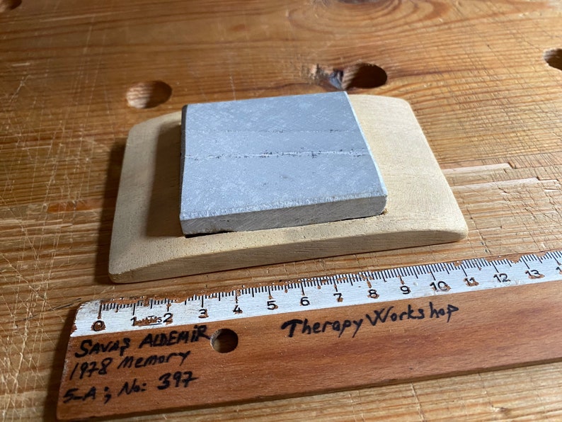 May include: A gray square stone resting on a wooden base. The base is curved and has a cutout for the stone. A wooden ruler with the text "Therapy Workshop" is visible in the foreground.