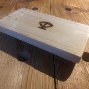 May include: A light wood block with a simple design burned into the surface. The design is a stylized image of a scale.
