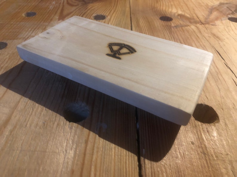 May include: A light wood block with a simple design burned into the surface. The design is a stylized image of a scale.