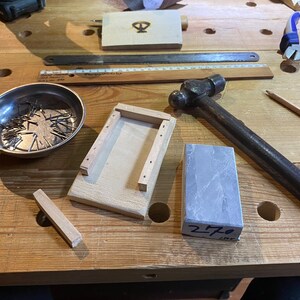 Peut inclure: Un &eacute;tabli en bois avec une vari&eacute;t&eacute; d'outils et de mat&eacute;riaux, dont un marteau, un bol en m&eacute;tal avec des clous, une bo&icirc;te en bois, une pierre &agrave; aiguiser et une r&egrave;gle. La pierre est &eacute;tiquet&eacute;e "270".