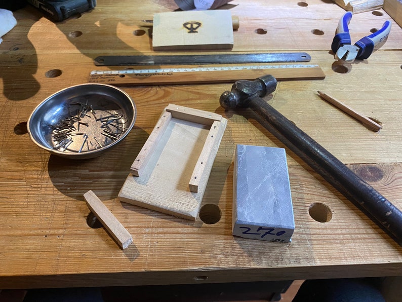 Peut inclure: Un &eacute;tabli en bois avec une vari&eacute;t&eacute; d'outils et de mat&eacute;riaux, dont un marteau, un bol en m&eacute;tal avec des clous, une bo&icirc;te en bois, une pierre &agrave; aiguiser et une r&egrave;gle. La pierre est &eacute;tiquet&eacute;e "270".