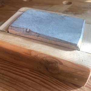 Puede incluir: Una piedra de afilar rectangular gris est&aacute; colocada en un marco de madera. El marco est&aacute; hecho de madera clara con un acabado natural.