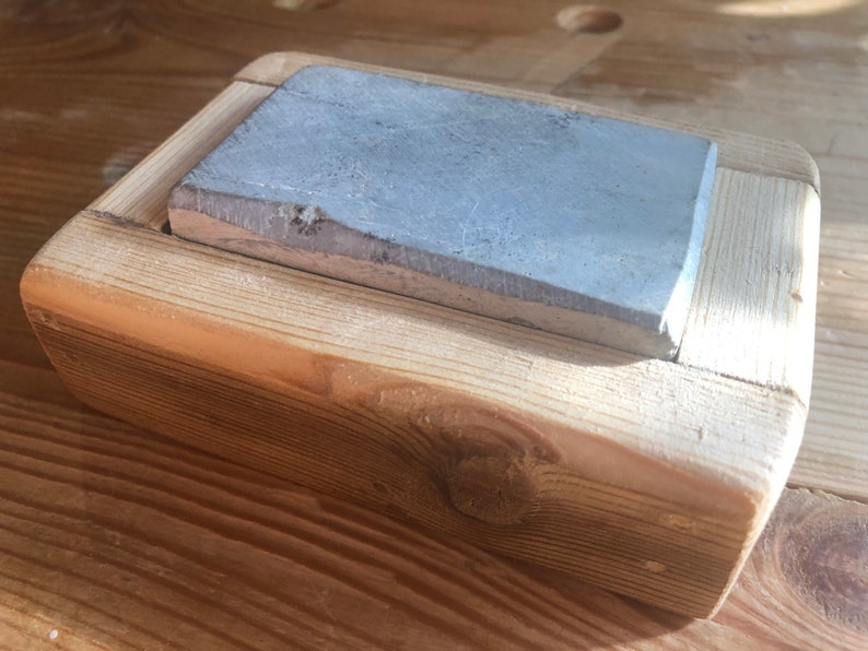 Puede incluir: Una piedra de afilar rectangular gris est&aacute; colocada en un marco de madera. El marco est&aacute; hecho de madera clara con un acabado natural.