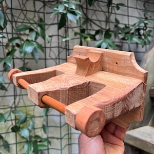 May include: A handcrafted wooden toilet paper holder. The holder is made of light brown wood with a natural finish. It features a horizontal dowel for the toilet paper roll and a decorative top piece. The holder is designed to be mounted on a wall.