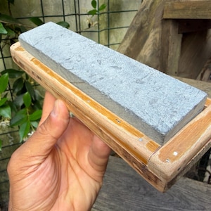 Op de afbeelding: Een rechthoekige grijze slijpsteen in een lichtbruine houten basis. De steen heeft een getextureerd, gevlekt oppervlak en is in het houten frame bevestigd met kleine metalen pinnen langs de randen, wat zorgt voor een stabiel slijphulpmiddel.