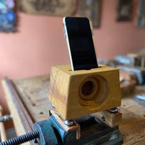 Peut inclure: Un support de téléphone en bois avec un haut-parleur intégré, serré dans un étau. Le support est en bois brun clair et a une forme rectangulaire. Un smartphone noir est placé dans le support.