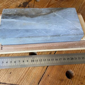 Puede incluir: Una piedra de afilar gris con una base de madera. La piedra mide aproximadamente 18 cm de largo y 5 cm de ancho. Una regla se coloca debajo de la base para la escala.
