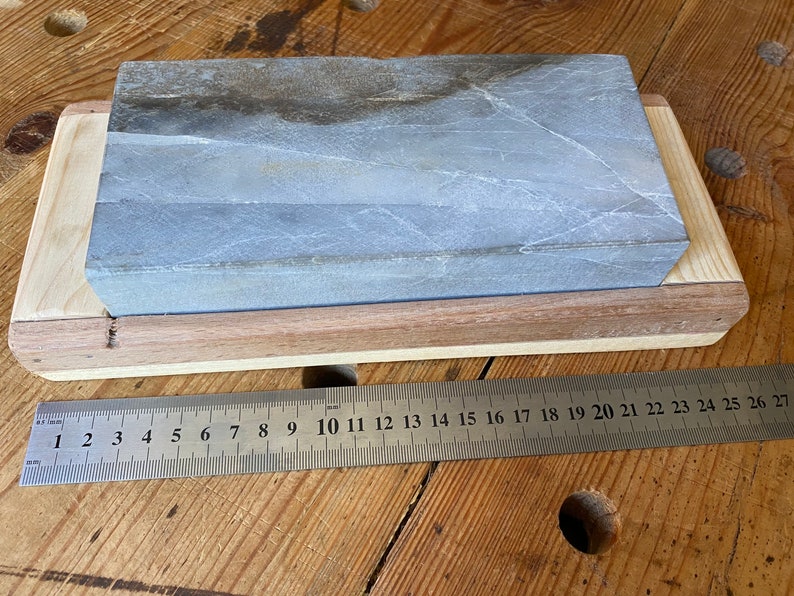 Puede incluir: Una piedra de afilar gris con una base de madera. La piedra mide aproximadamente 18 cm de largo y 5 cm de ancho. Una regla se coloca debajo de la base para la escala.
