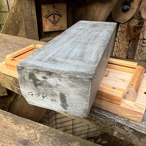 May include: A rectangular gray stone block with a textured surface and visible white markings, featuring the handwritten text 1715 gr. on its front face, resting on a light-colored wooden frame with two parallel rails and small metal fasteners.