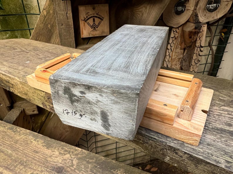 May include: A rectangular gray stone block with a textured surface and visible white markings, featuring the handwritten text 1715 gr. on its front face, resting on a light-colored wooden frame with two parallel rails and small metal fasteners.