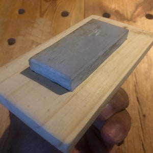 May include: A grey rectangular sharpening stone mounted on a wooden base. The stone is made of a fine-grained material and is ideal for sharpening knives.