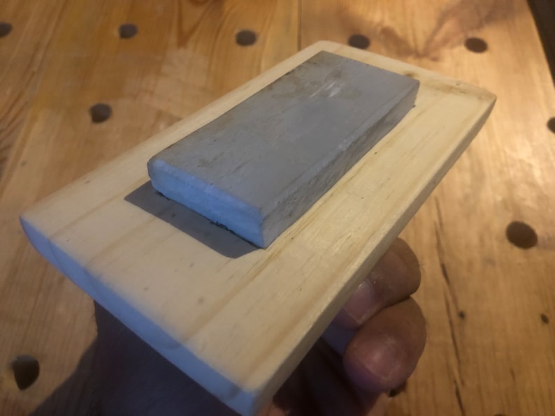 May include: A grey rectangular sharpening stone mounted on a wooden base. The stone is made of a fine-grained material and is ideal for sharpening knives.