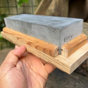 May include: A rectangular gray sharpening stone mounted on a light-colored wooden base with two side rails. The stone features a handwritten inscription on its side that reads 1715 gr, indicating its weight in grams.