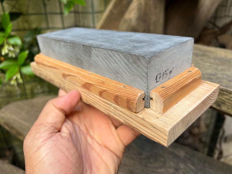 May include: A rectangular gray sharpening stone mounted on a light-colored wooden base with two side rails. The stone features a handwritten inscription on its side that reads 1715 gr, indicating its weight in grams.