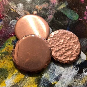 May include: Three copper discs, one smooth, one hammered, and one with a textured surface. The discs are arranged on a colourful background.