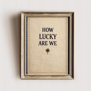 May include: Framed artwork with the text "HOW LUCKY ARE WE" in navy blue. A small green four-leaf clover is below the text. The artwork is on a beige background with a stitched border and a silver frame.